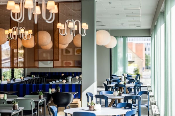 Un intérieur de restaurant moderne, doté de plusieurs espaces de restauration, notamment des tables et des banquettes, ainsi que de luminaires suspendus.