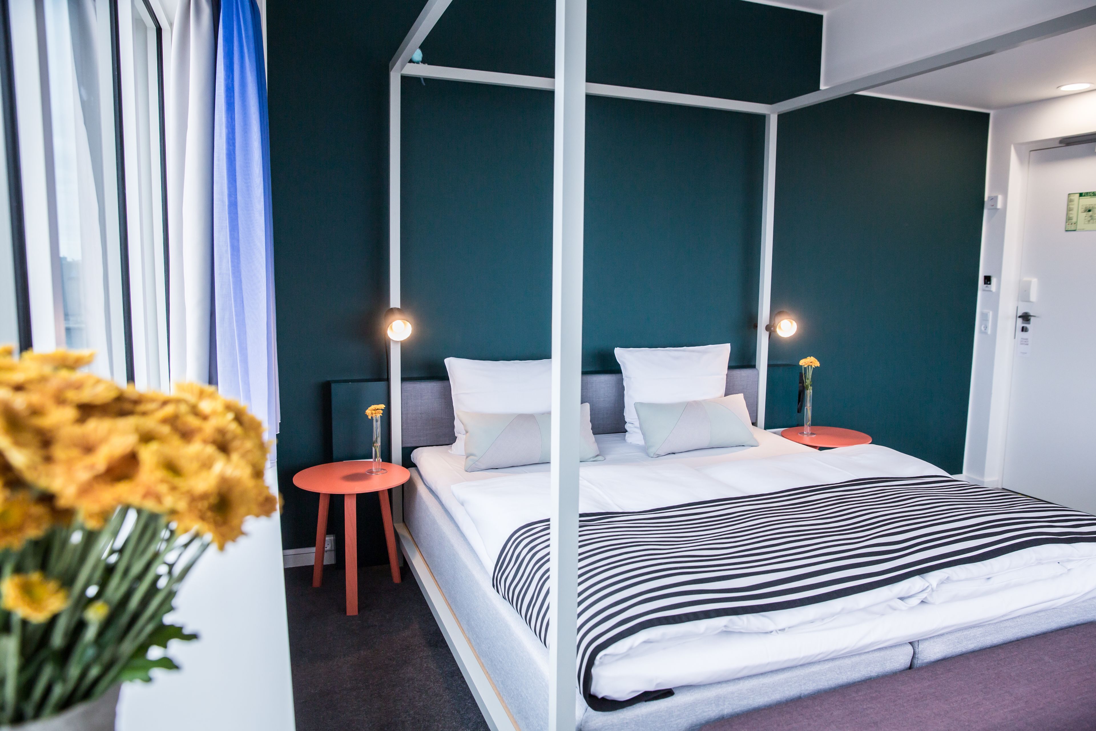 A four-poster bed with white bedding and a striped blanket, two bedside tables with lamps, a small stool, and a vase with flowers.