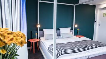 A four-poster bed with white bedding and a striped blanket, two bedside tables with lamps, a small stool, and a vase with flowers.