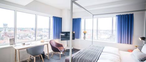 A modern hotel room with a large bed, a desk, and a chair. There is a TV on the desk and a window with curtains.