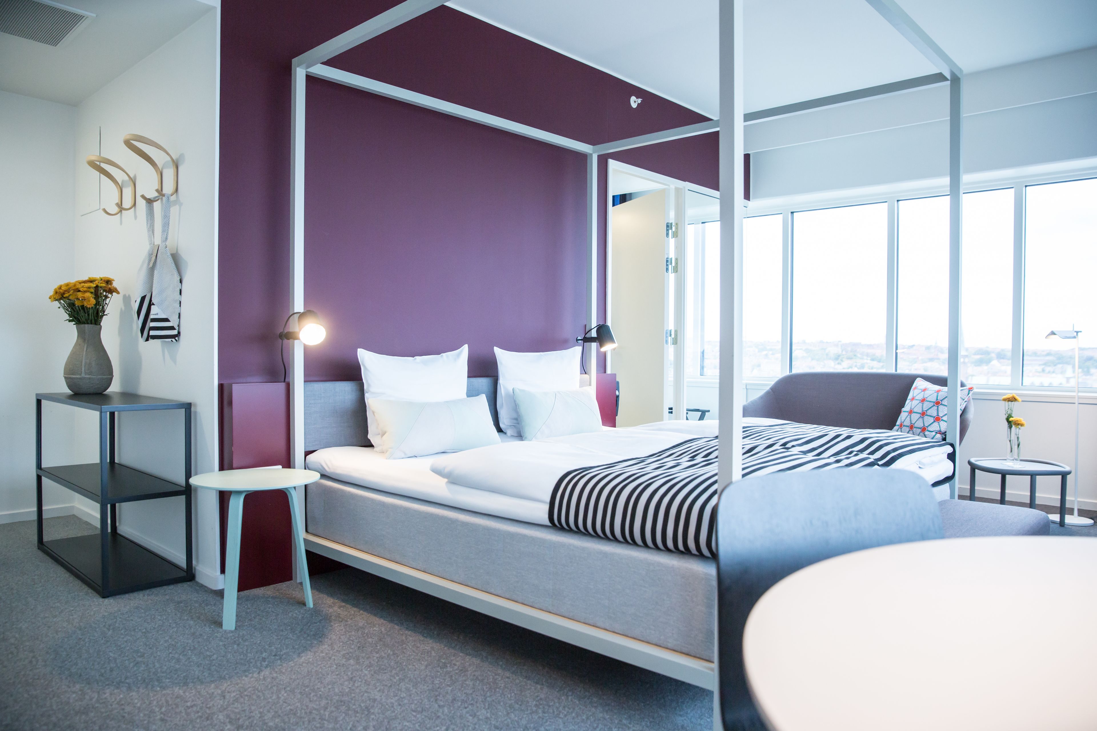 A modern bedroom with a four-poster bed, a small side table, a chair, and a view of the city through large windows.