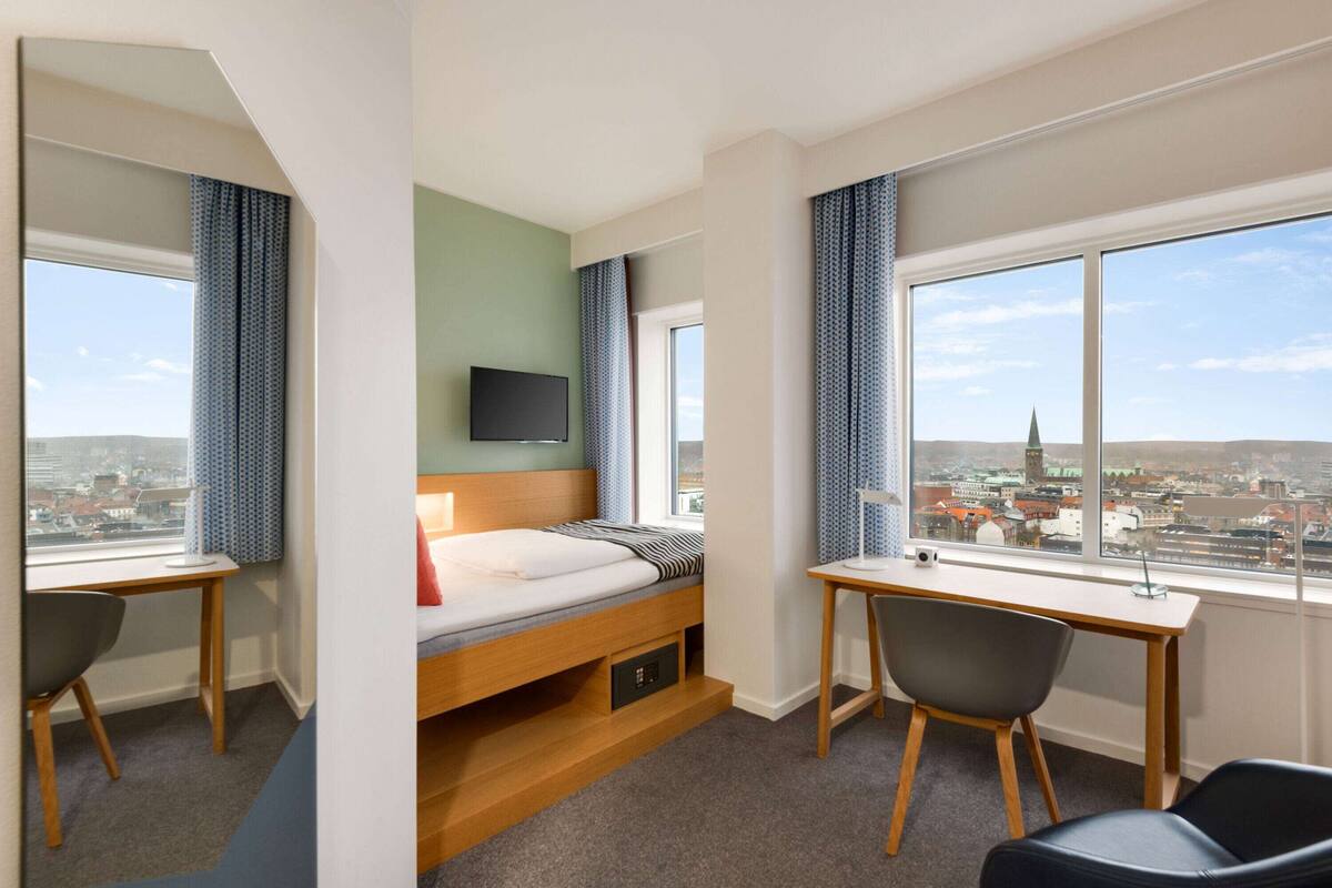 Een hotelkamer met een bed, bureau en stoelen, een televisie en grote ramen met uitzicht op de stad.