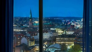 Een stadsgezicht dat te zien is door een raam, met een opvallende kerktoren en een dichtbebouwd stedelijk gebied met verlichte gebouwen.
