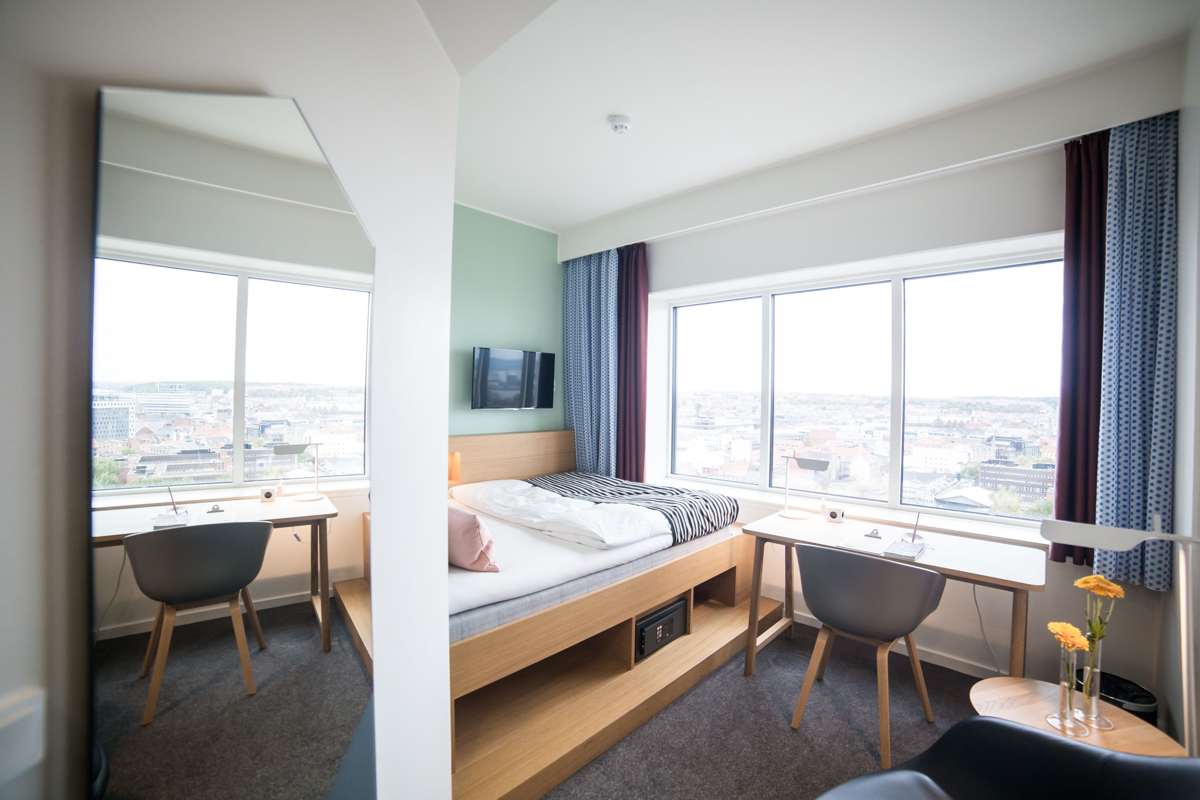 A modern hotel room with a bed, desk, and chairs, large windows with a city view, and a flat-screen TV on the wall.