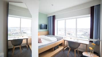 A modern hotel room with a bed, desk, and chairs, large windows with a city view, and a flat-screen TV on the wall.