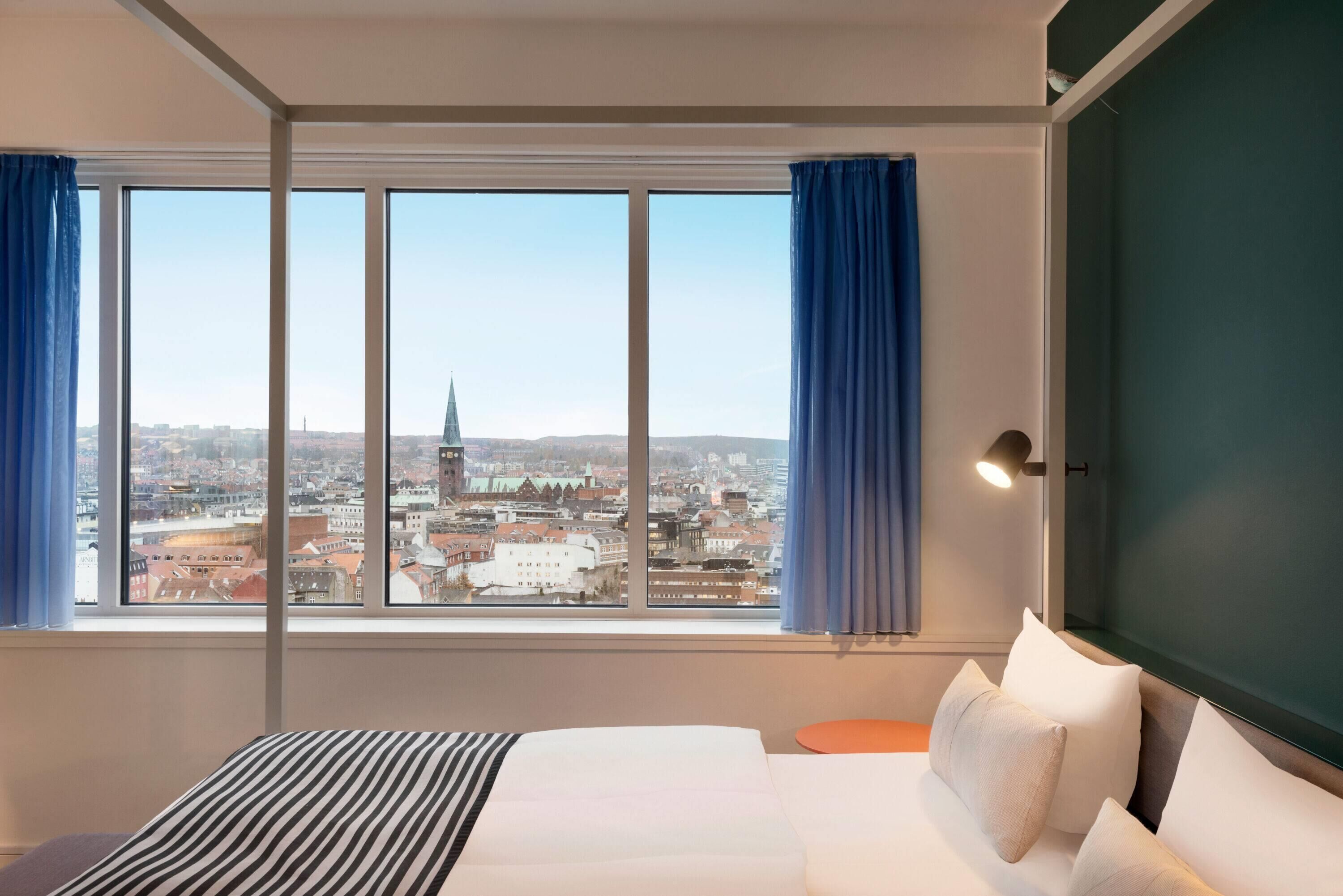 Et hotelværelse med et stort vindue, en seng, et sengebord og en lampe.