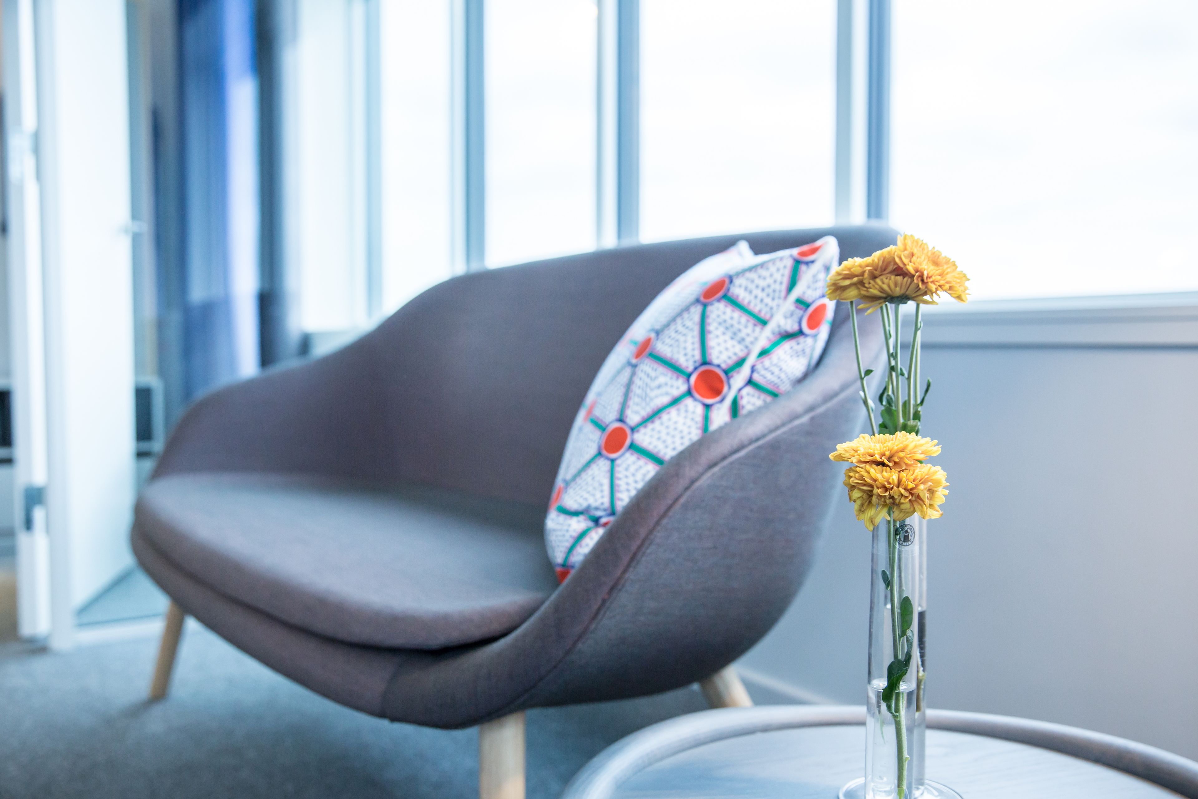 Un fauteuil moderne agrémenté d’un coussin aux motifs géométriques, un vase transparent rempli de fleurs et une vue sur un bâtiment à travers de grandes fenêtres.