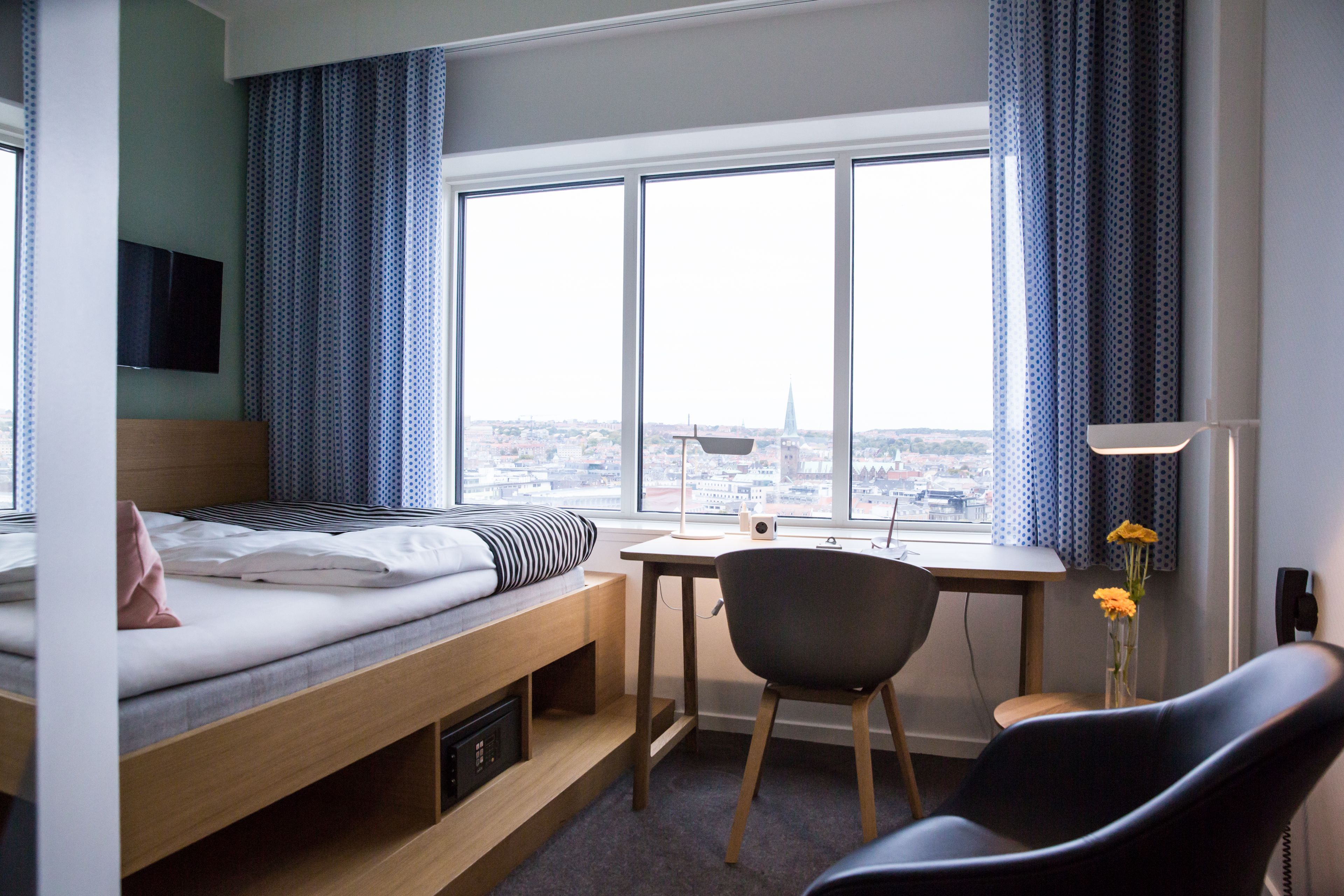Een hotelkamer met een bed, bureau, stoel en groot raam met uitzicht op de stad.