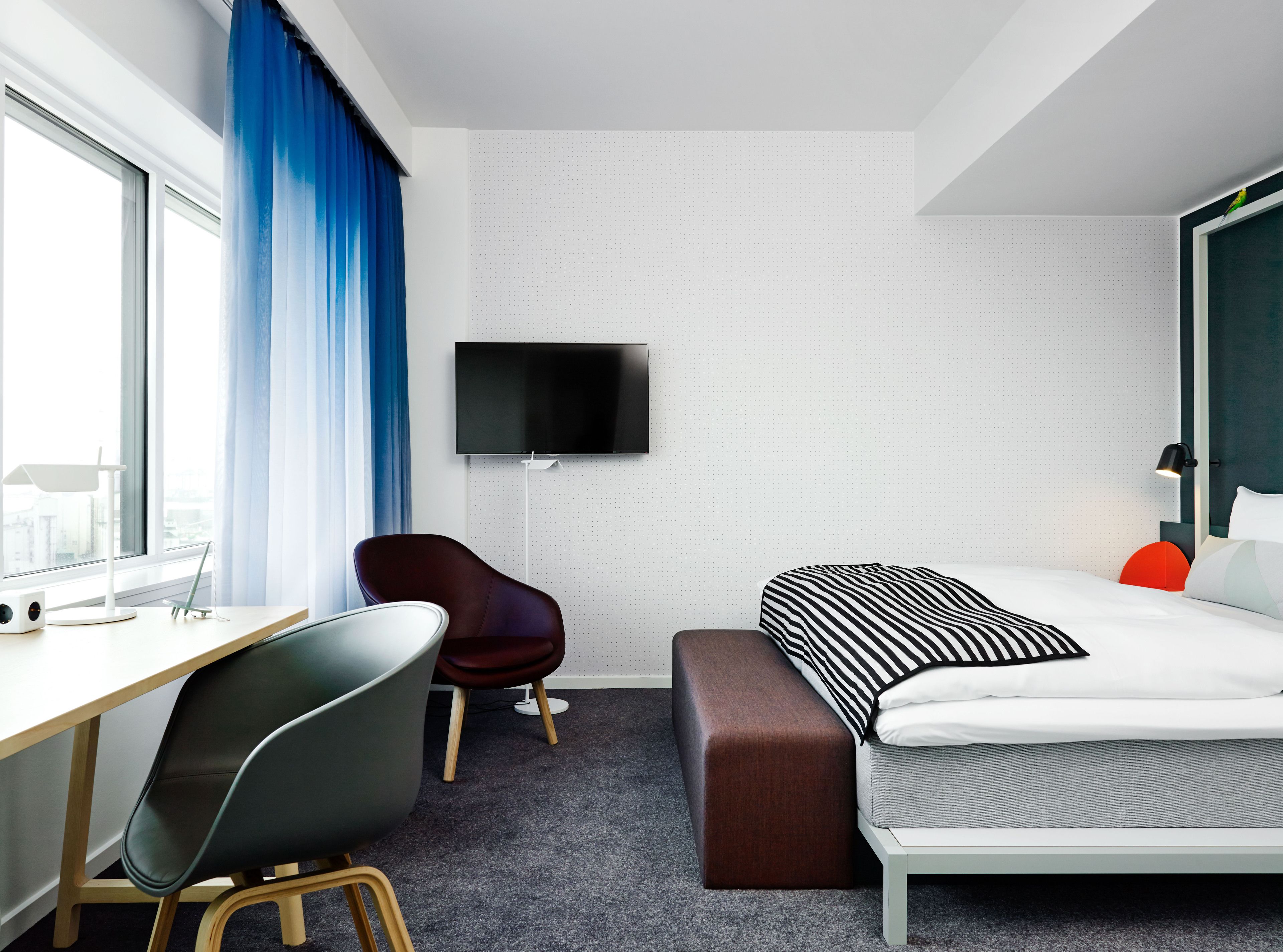 A modern hotel room with a desk, chair, bed, and a TV mounted on the wall.