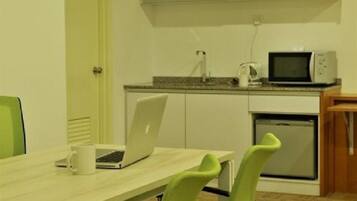 A small kitchenette with a microwave, a coffee maker, and a laptop on a table. There are two chairs and a door leading to another room.