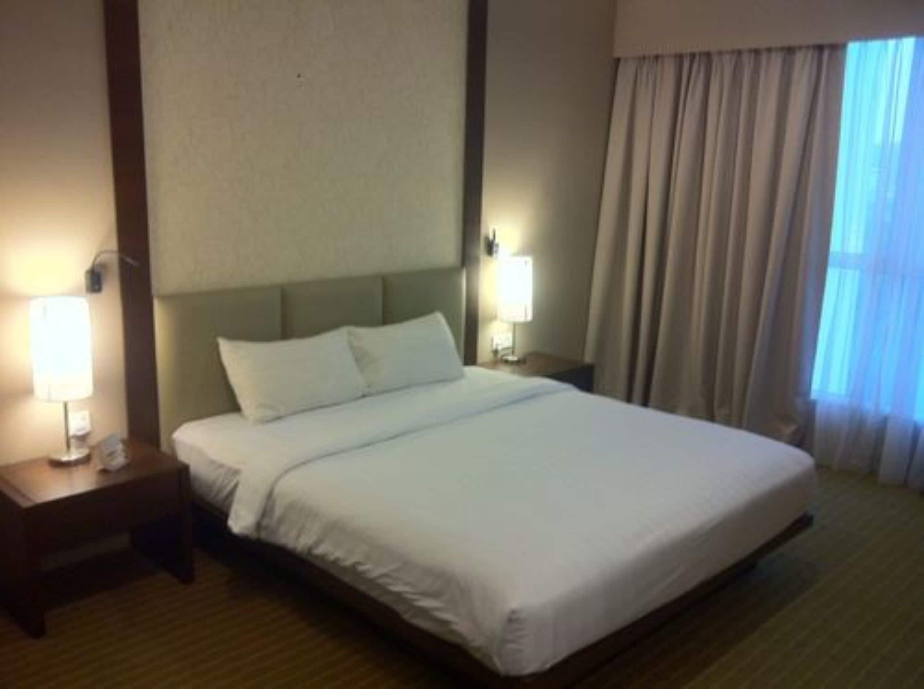 A hotel room with a large bed, two bedside tables with lamps, a headboard, and a window with curtains.