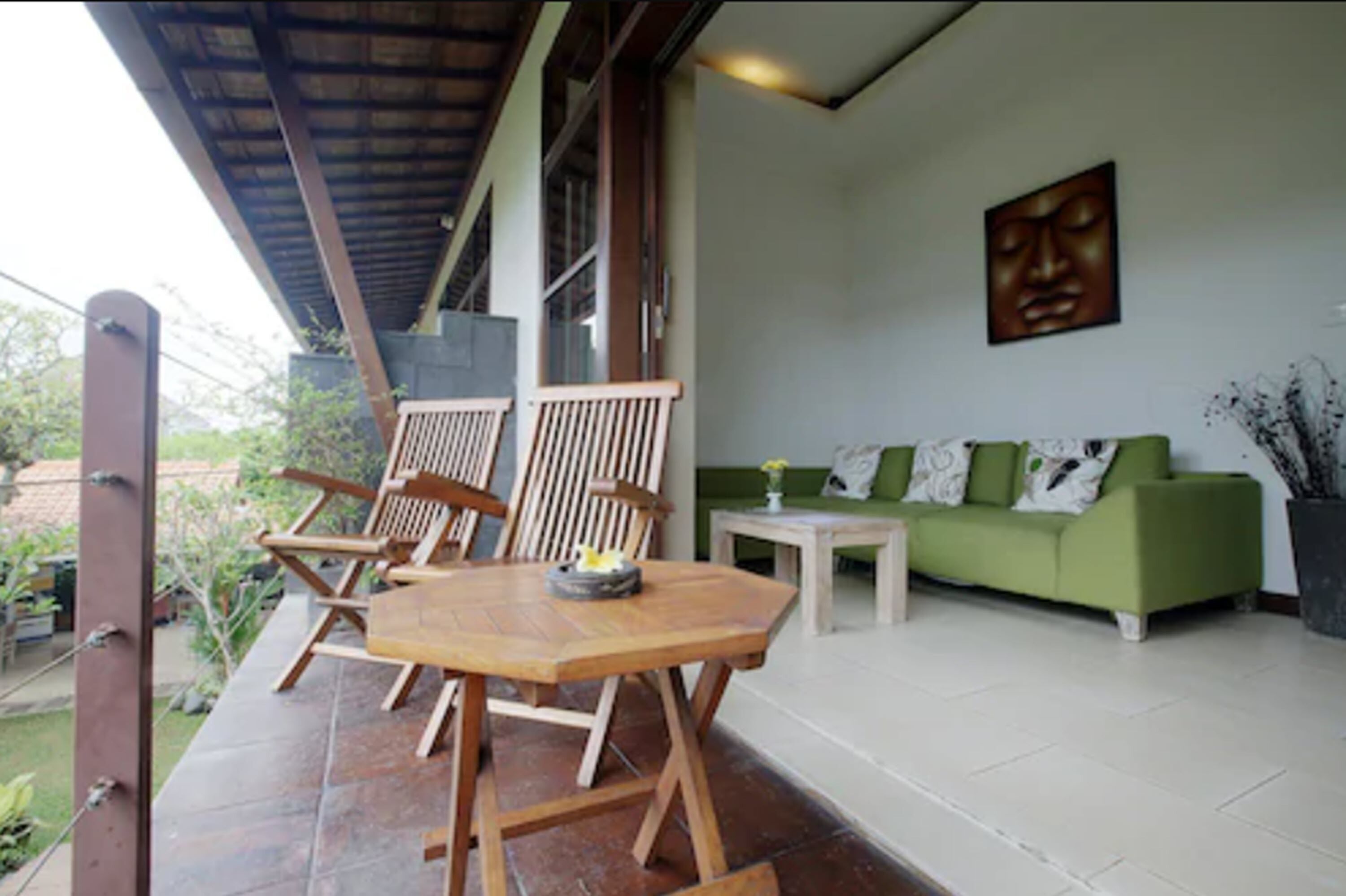 A covered outdoor seating area with wooden chairs and a table, a green sofa, and a wall-mounted artwork.