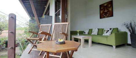 A covered outdoor seating area with wooden chairs and a table, a green sofa, and a wall-mounted artwork.