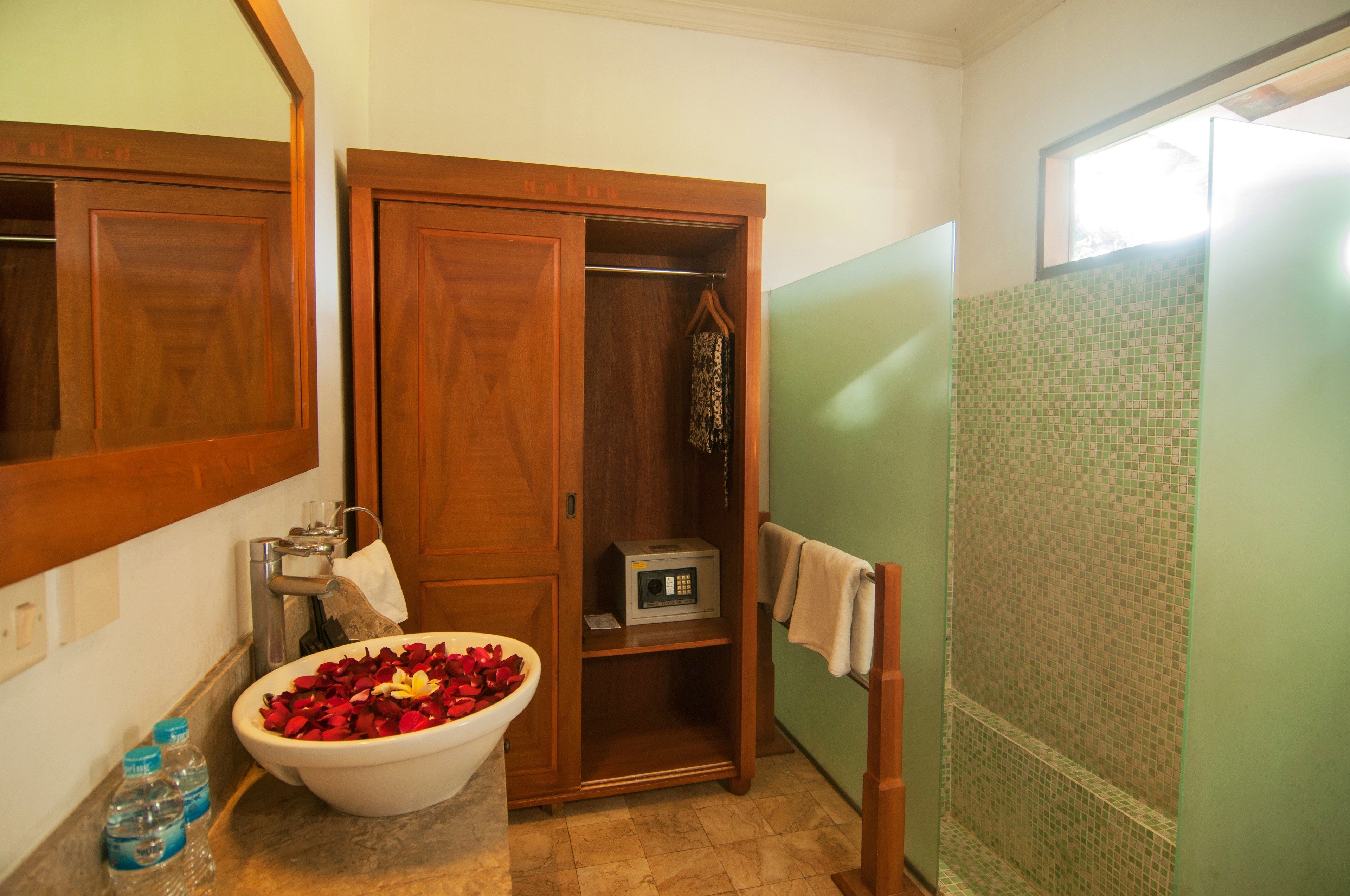 A bathroom with a wooden vanity, a bowl of rose petals, a microwave, and a shower with green mosaic tiles.