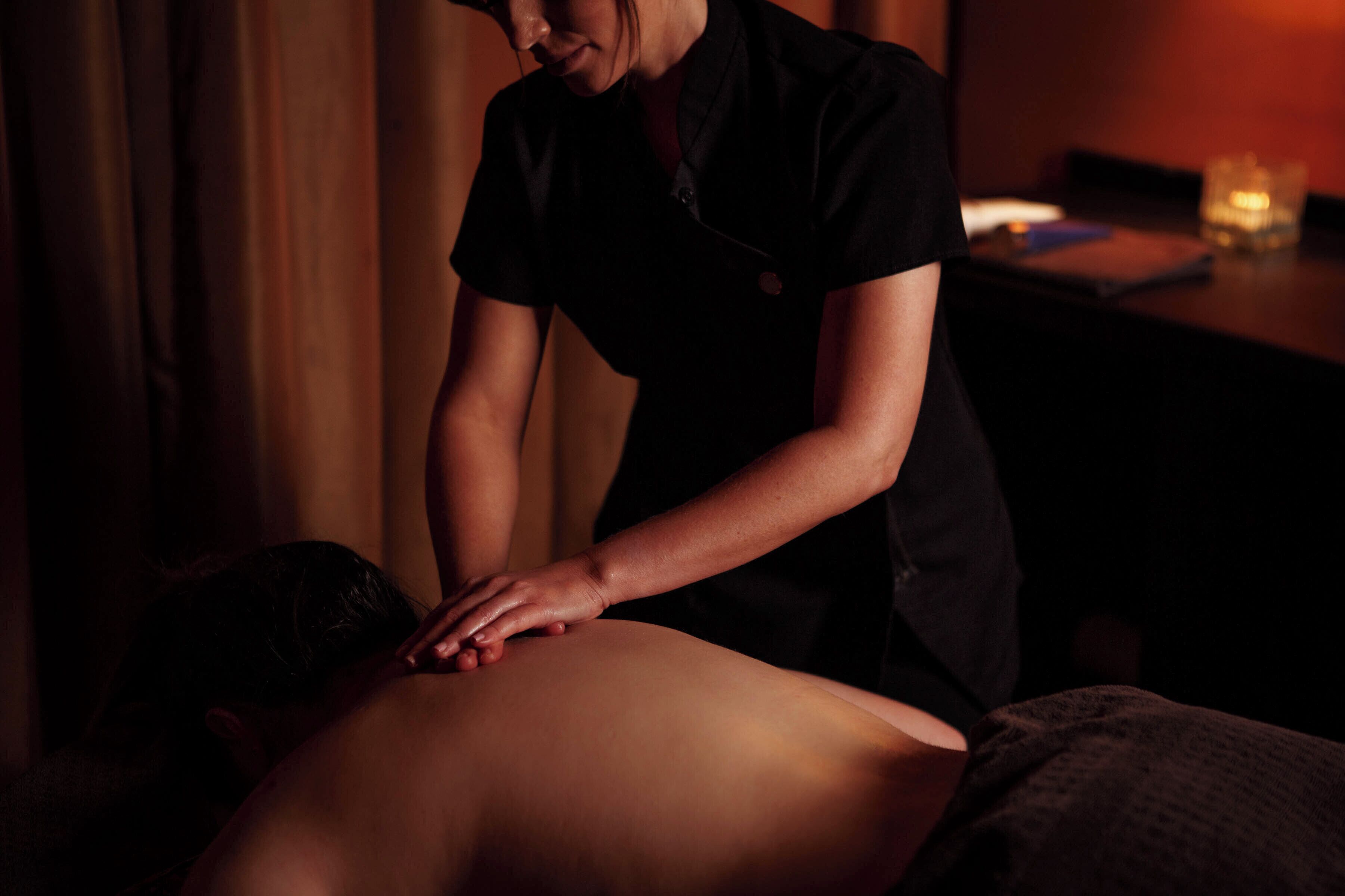 Sesión de masoterapia en una sala con poca luz, con una camilla, un cliente boca abajo y un terapeuta a su lado.