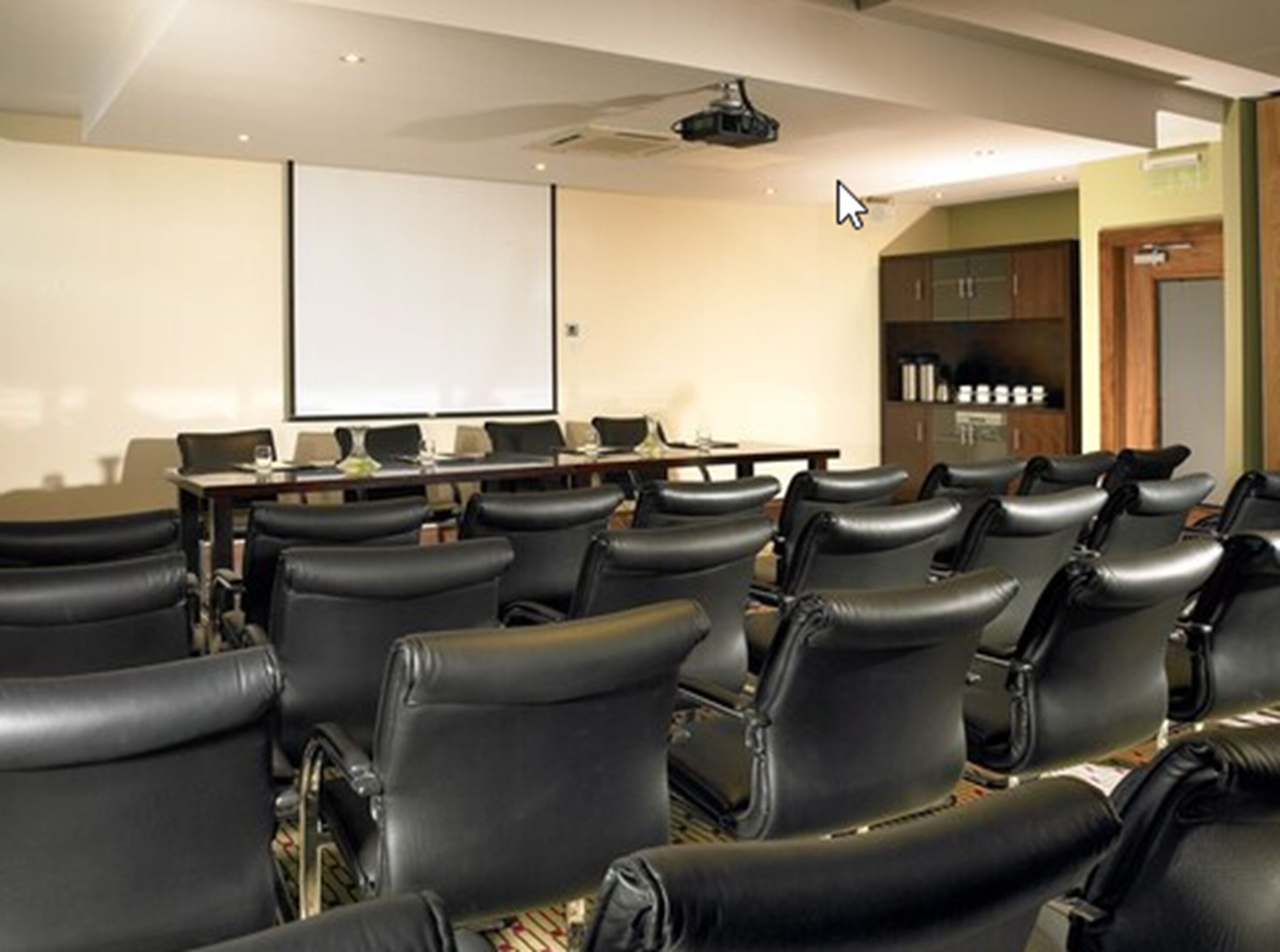 A conference room with rows of black leather chairs, a projector screen, a table with chairs, and a bookshelf with binders.