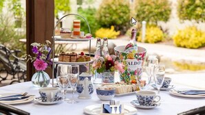 Une table dressée pour un service à thé, avec un présentoir à étages rempli de desserts, des arrangements floraux et une bouteille de vin Aldo.