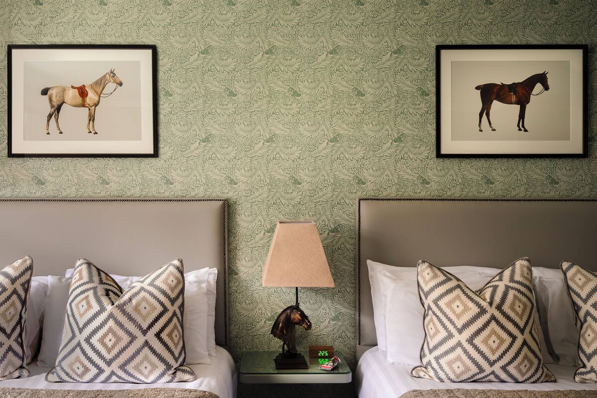 Une chambre d’hôtel avec deux lits, des coussins à motifs, une table de chevet avec une lampe et des tableaux représentant des chevaux accrochés au mur.