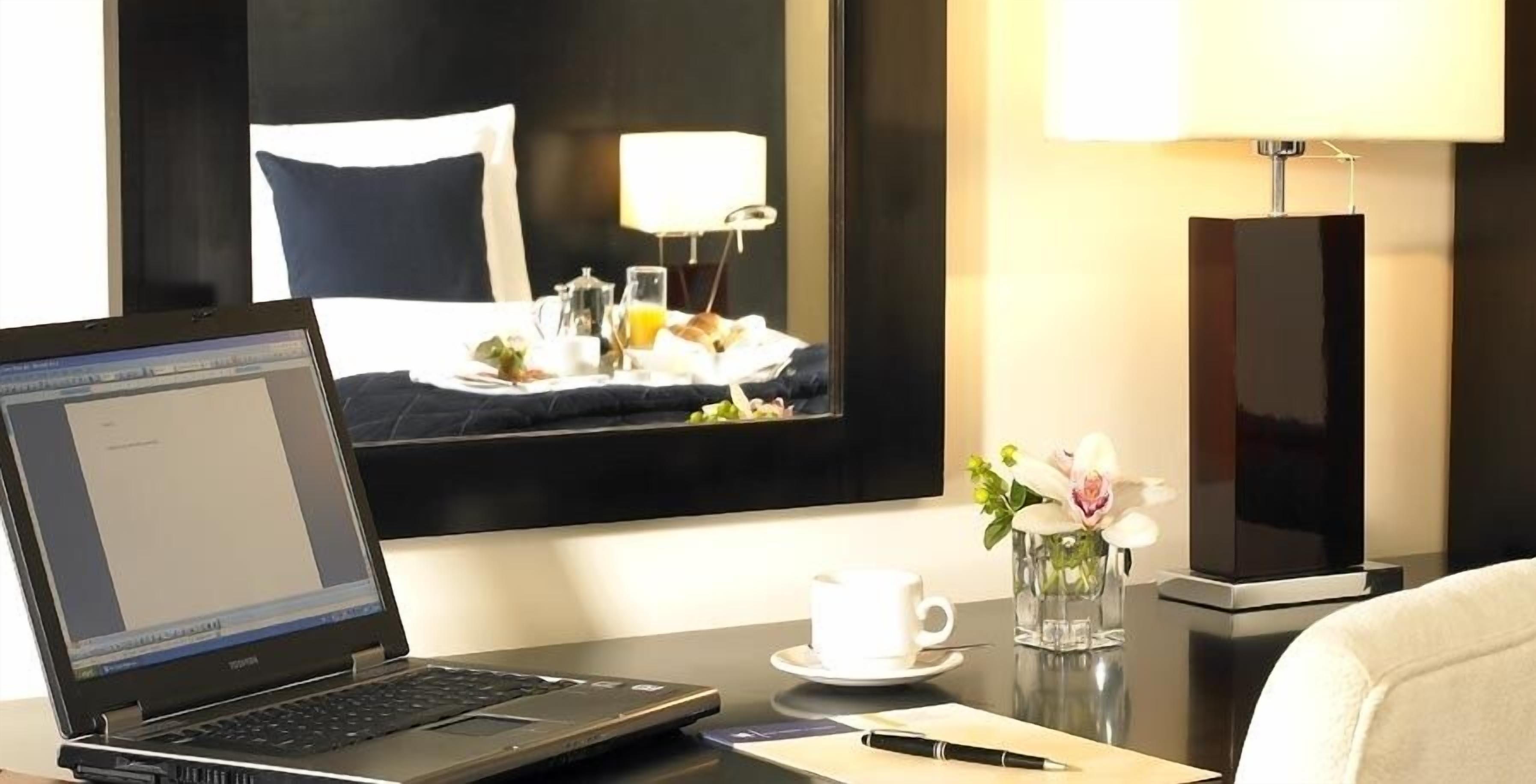 Une chambre d’hôtel avec un ordinateur portable sur un bureau, une tasse, un vase avec des fleurs et un lit avec une tête de lit noire et blanche.