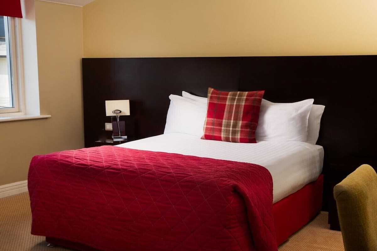 Une chambre d’hôtel avec un grand lit, une couvre-lit matelassée rouge et blanche, une tête de lit noire, un coussin à carreaux rouges, un coussin blanc, une petite table de chevet avec une lampe et une chaise jaune.