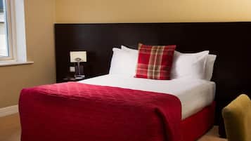 Hotelkamer met een groot bed, een rood-wit geborduurde sprei, een zwart hoofdeinde, een rood geruit kussen, een wit kussen, een klein nachtkastje met een lamp, en een gele stoel.