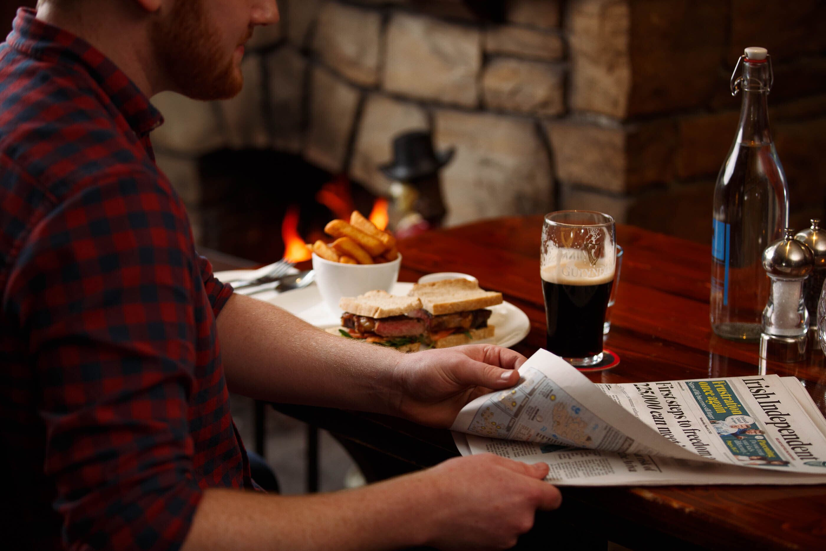 Una persona leyendo un periódico en una mesa, con un sándwich, papas fritas, una cerveza en un vaso y una botella.