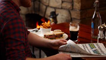Una persona leyendo un periódico en una mesa, con un sándwich, papas fritas, una cerveza en un vaso y una botella.