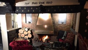 A cozy room with a fireplace, a red chair, a patterned armchair, and a sign above the fireplace that reads "Please Mind Your Head".
