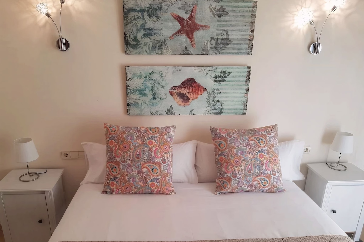 Un dormitorio con una cama, dos almohadas estampadas, una obra de arte con forma de estrella de mar y apliques de pared.