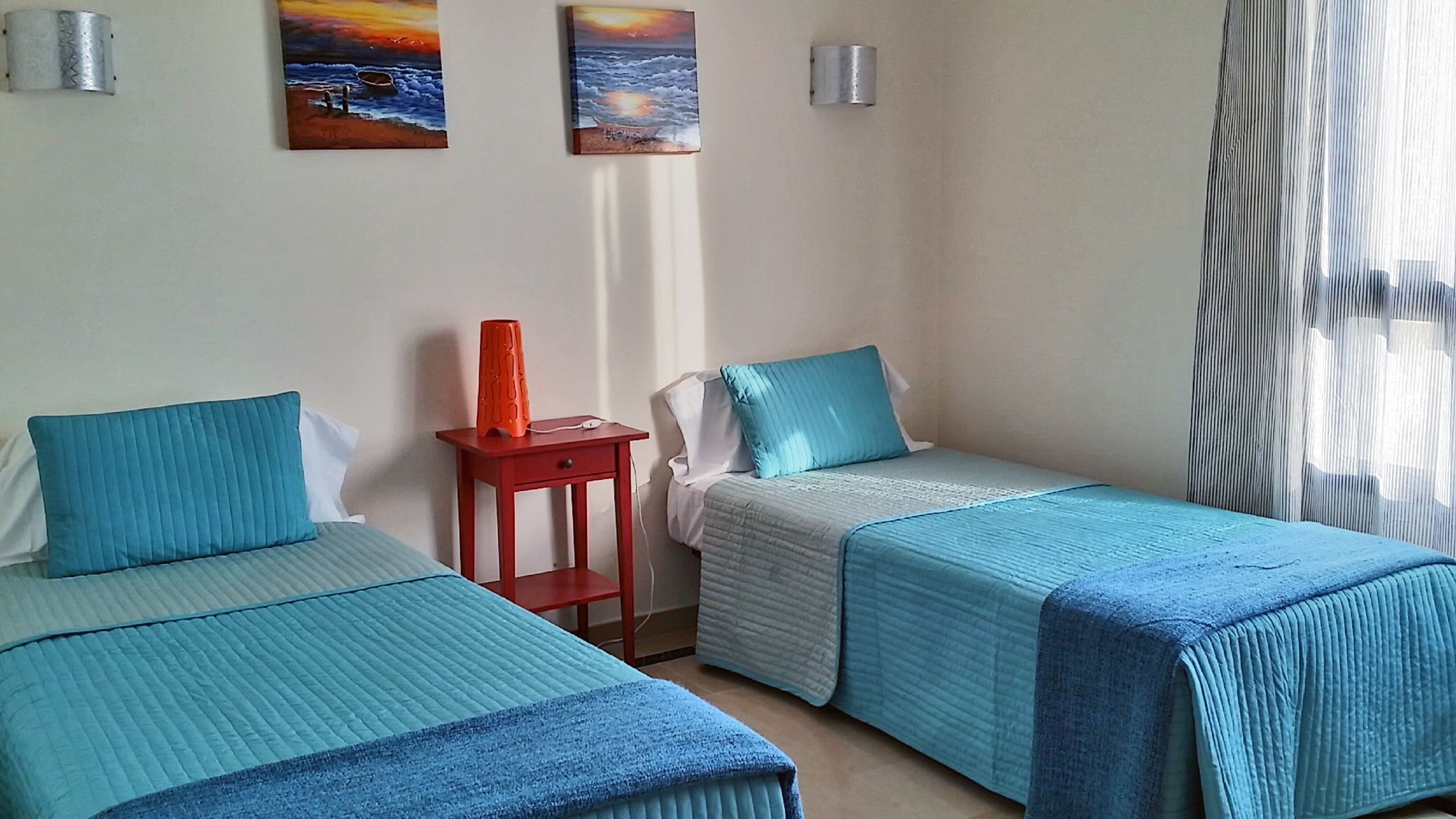 A hotel room with two beds, each with a blue bedspread and pillow, a small red table, and two wall-mounted lamps.