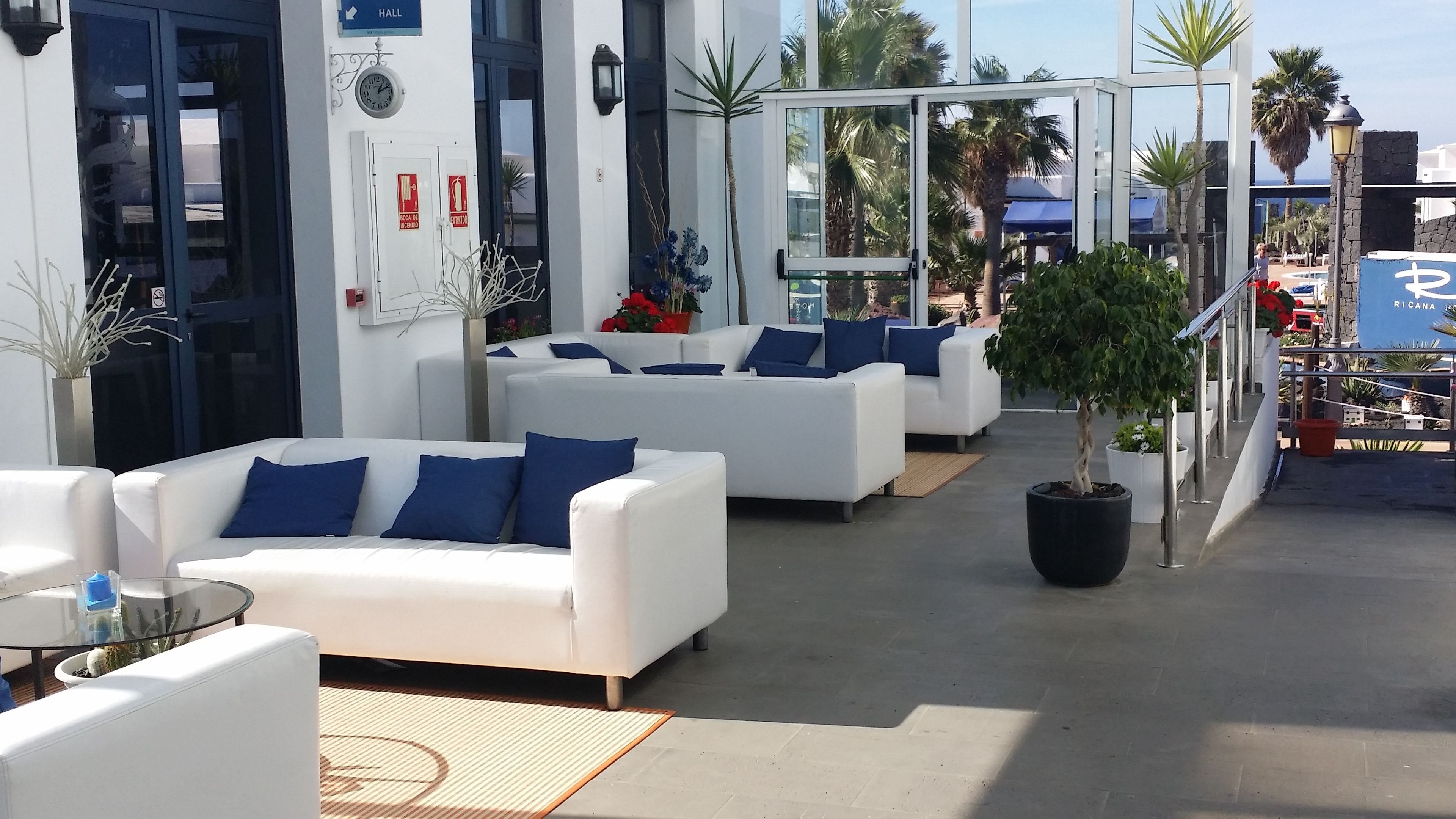 Un espace de détente moderne avec des canapés blancs et des coussins bleus, un arbre en pot et une porte vitrée donnant sur l’extérieur.