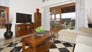 A living room with a TV, wooden furniture, a potted plant, and a view through a large window. - Villas Coral Deluxe (Yaiza)