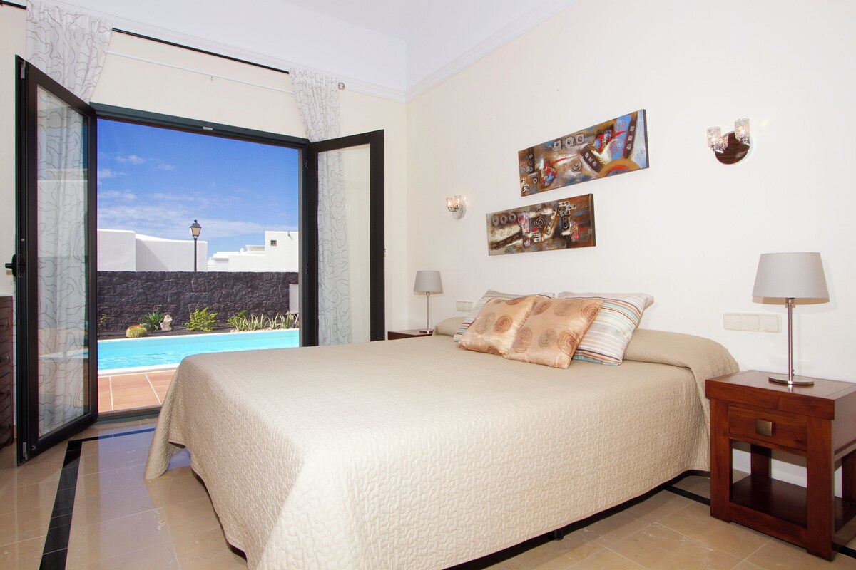 Une chambre avec un grand lit, une table de chevet, une lampe et une vue sur une piscine.