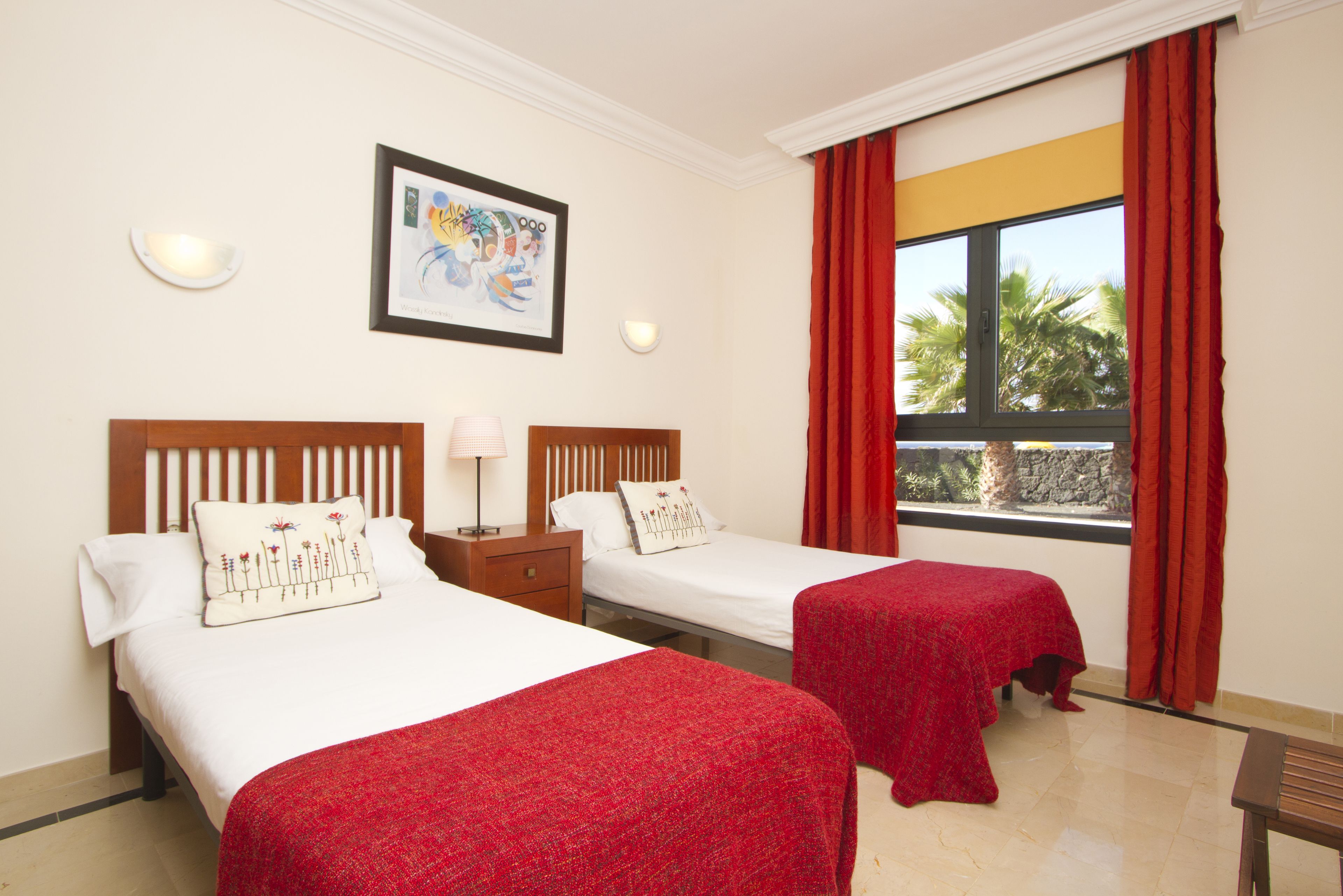 A hotel room with two beds, a large window with red curtains, a framed artwork on the wall, and a small table with a lamp.