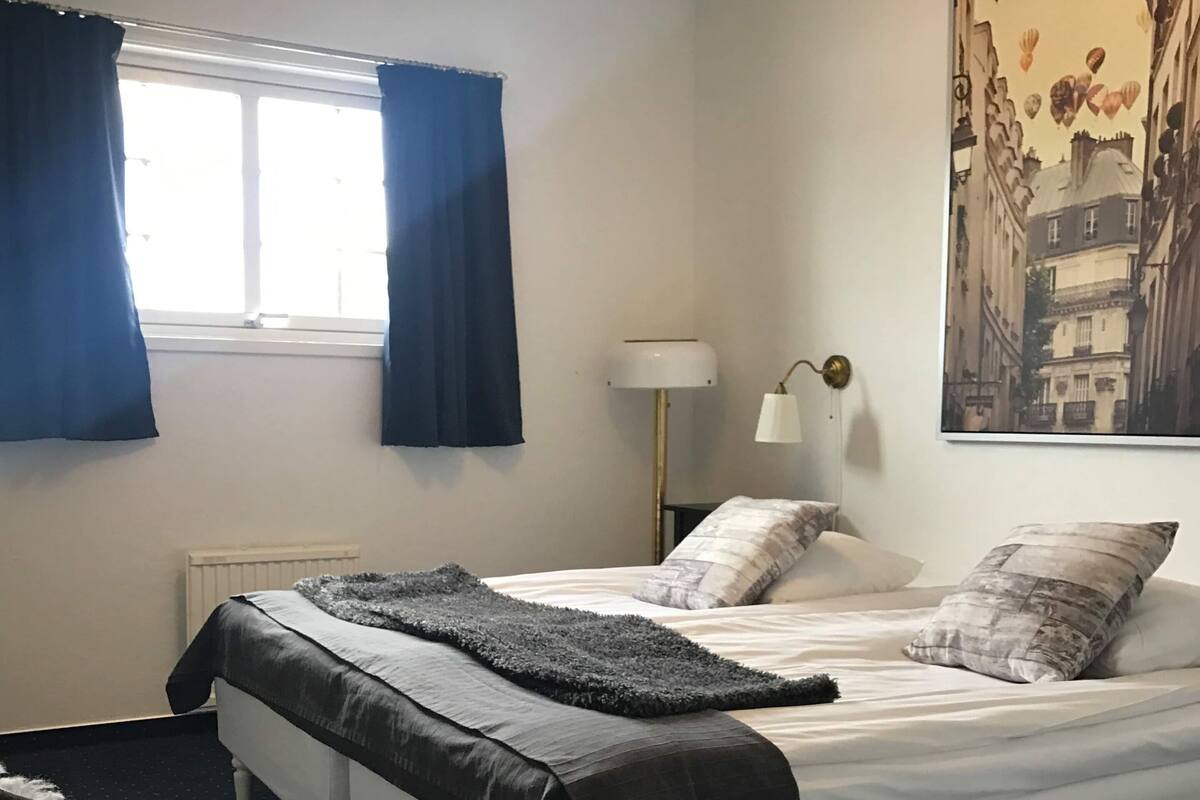 A bedroom with a bed, a chair, a window with curtains, a lamp, and a cityscape painting.