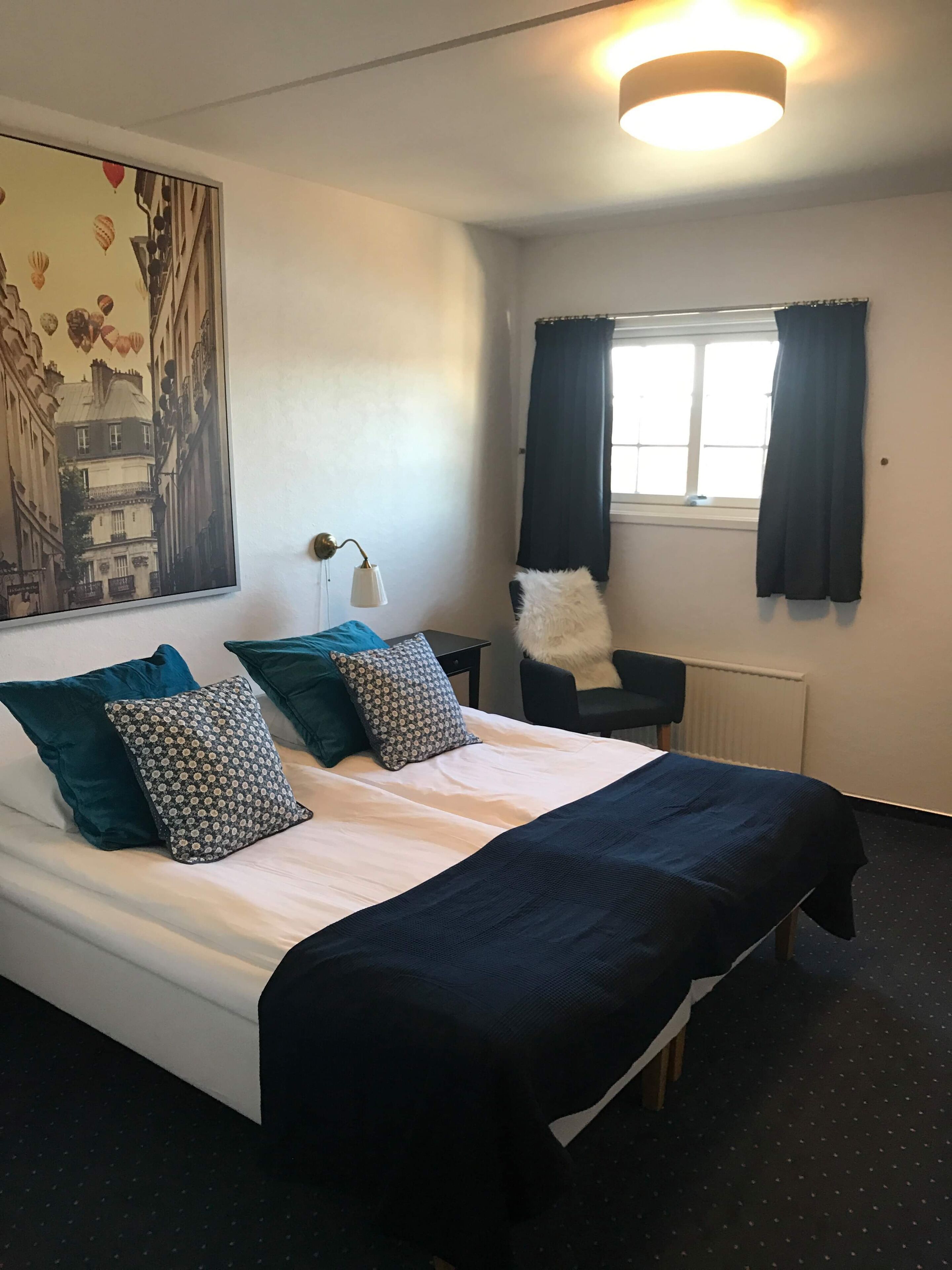 A neatly made bed with a white duvet and dark blue bedding, a black chair with a fluffy pillow, a lamp on a bedside table, and a large cityscape painting on the wall.