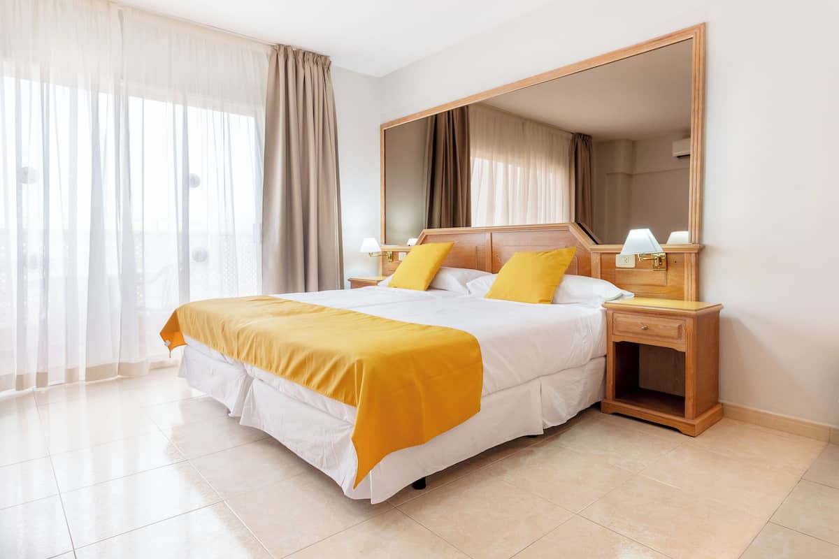 A hotel room with a large bed, a nightstand, a mirror, and sheer curtains.