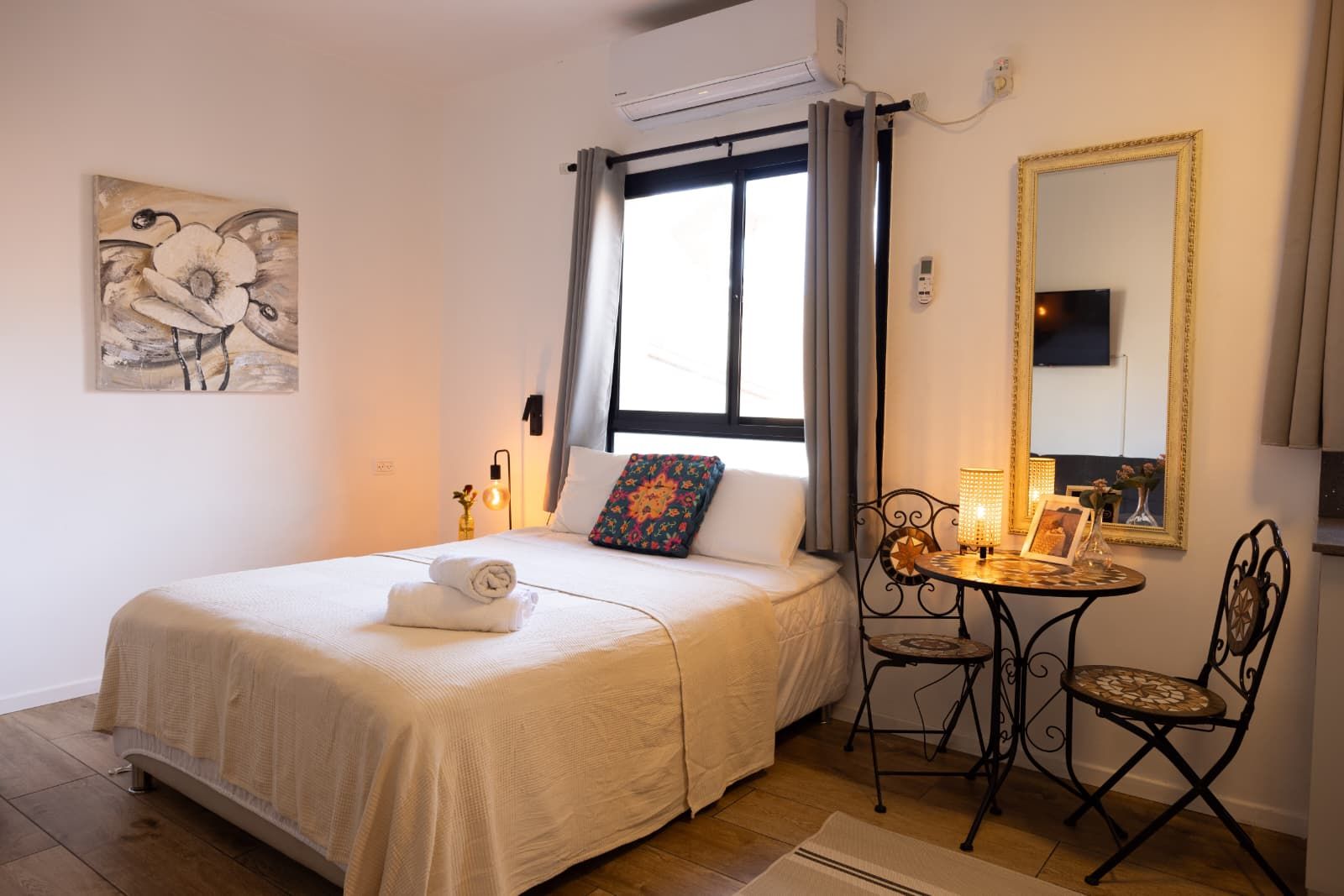 A hotel room with a bed, a small table with a lamp, a chair, a mirror, and a painting on the wall.