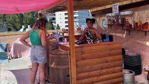 Een houten buitenbar met een roze luifel, een houten vat en kunstgras.