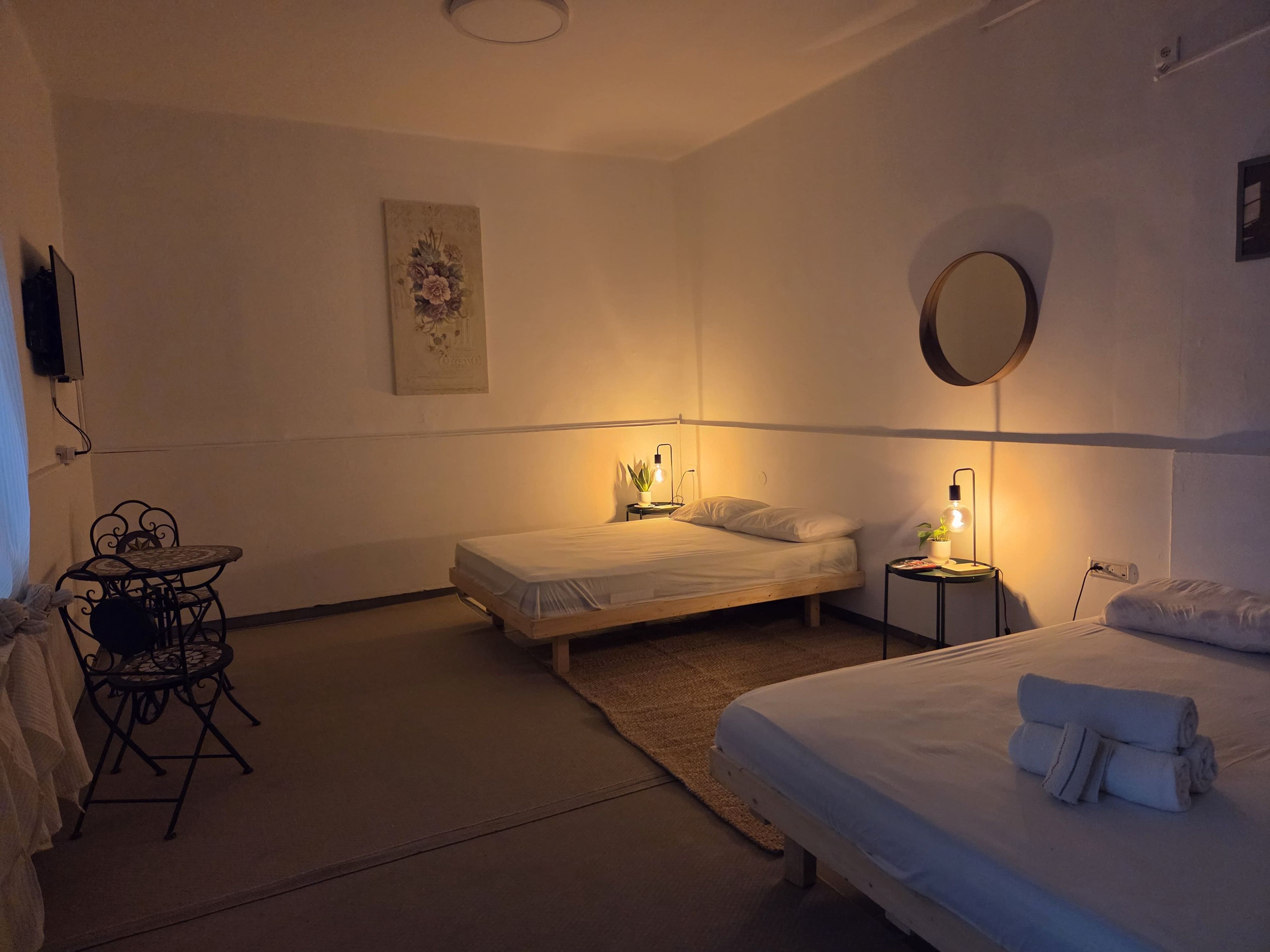 Een slaapkamer met twee bedden, een kleintje tafeltje met stoelen, een spiegel aan de muur, en één lichtbron.