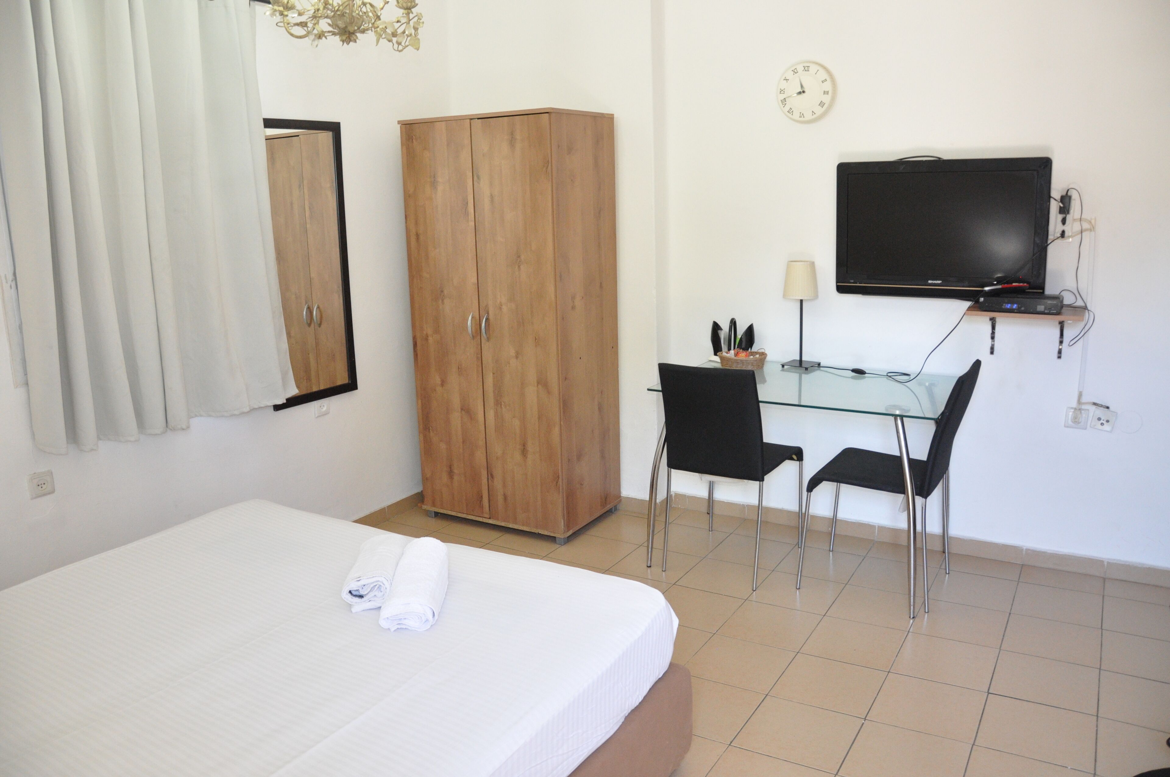 A bedroom with a bed, a wooden wardrobe, a desk with two chairs, a television, and a clock on the wall.