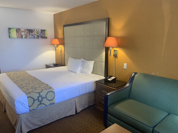 A hotel room with a large bed, two bedside lamps, a green armchair, and a small table. - Brentwood Inn & Suites (Hobbs)