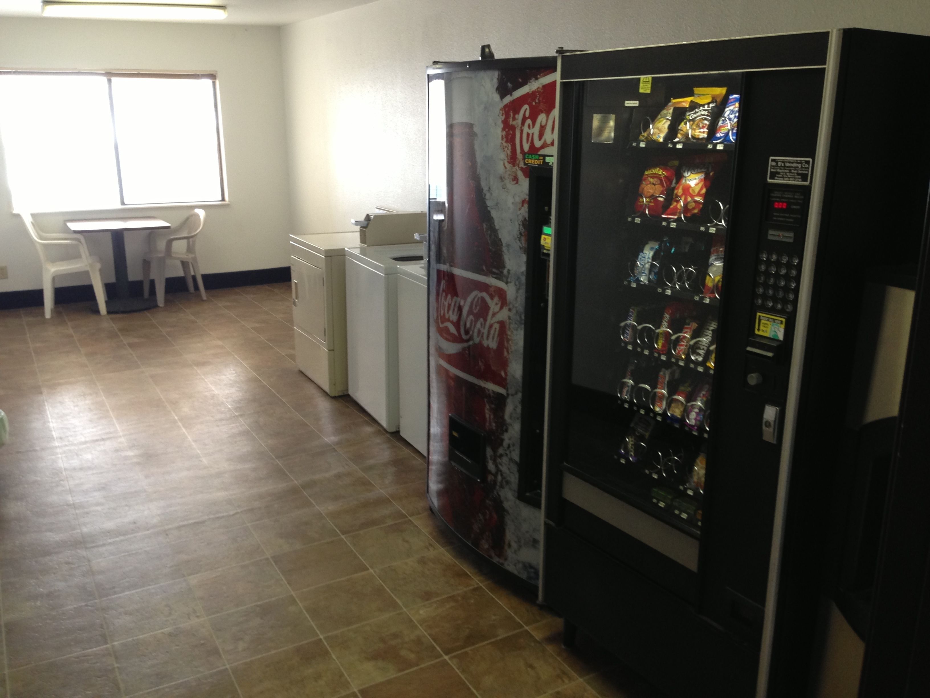 A vending machine with snacks and a Coca-Cola refrigerator in a room with a tiled floor and a window.