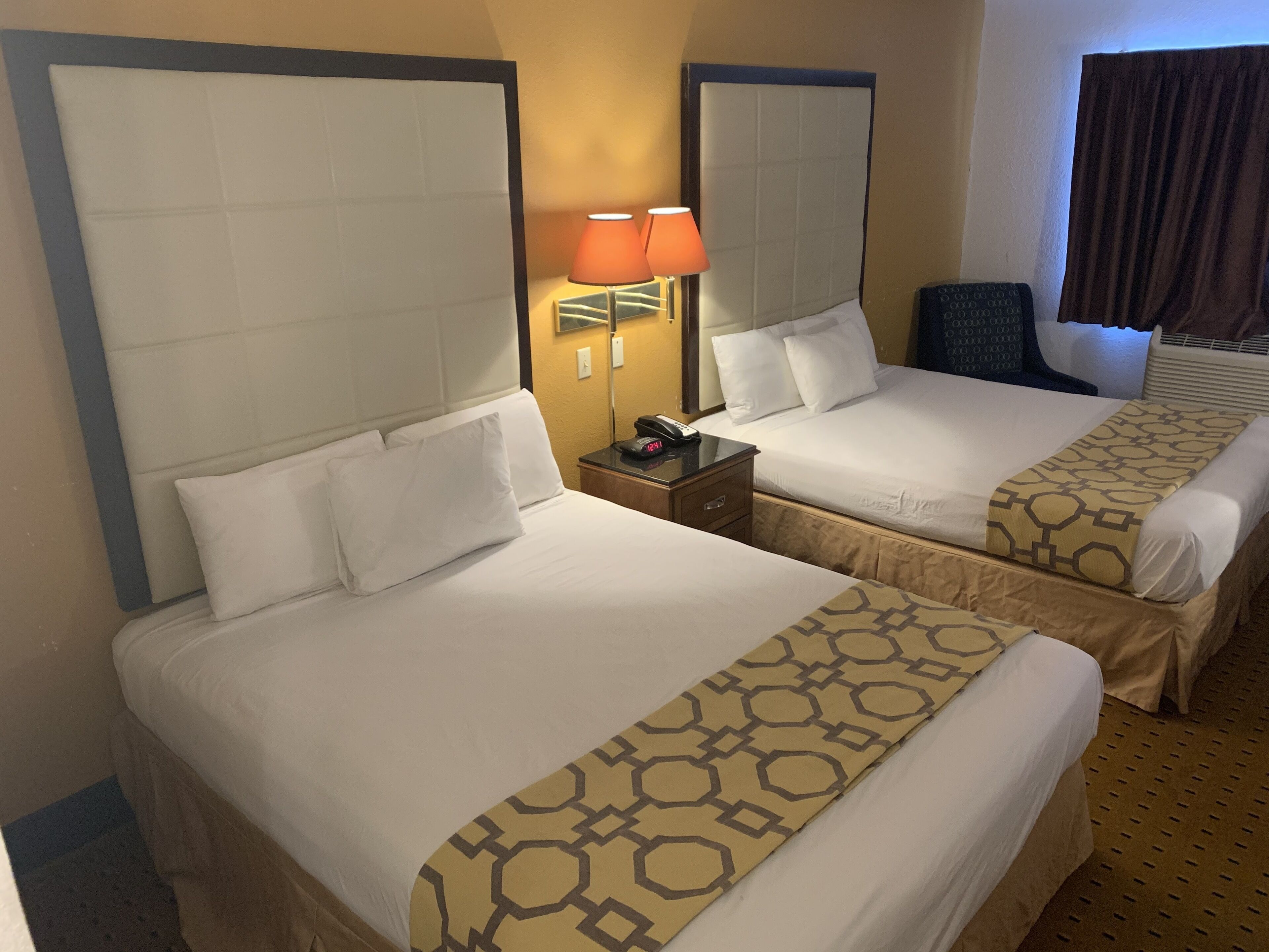 Two adjacent hotel beds with headboards, a bedside table with a lamp, and a window with curtains.