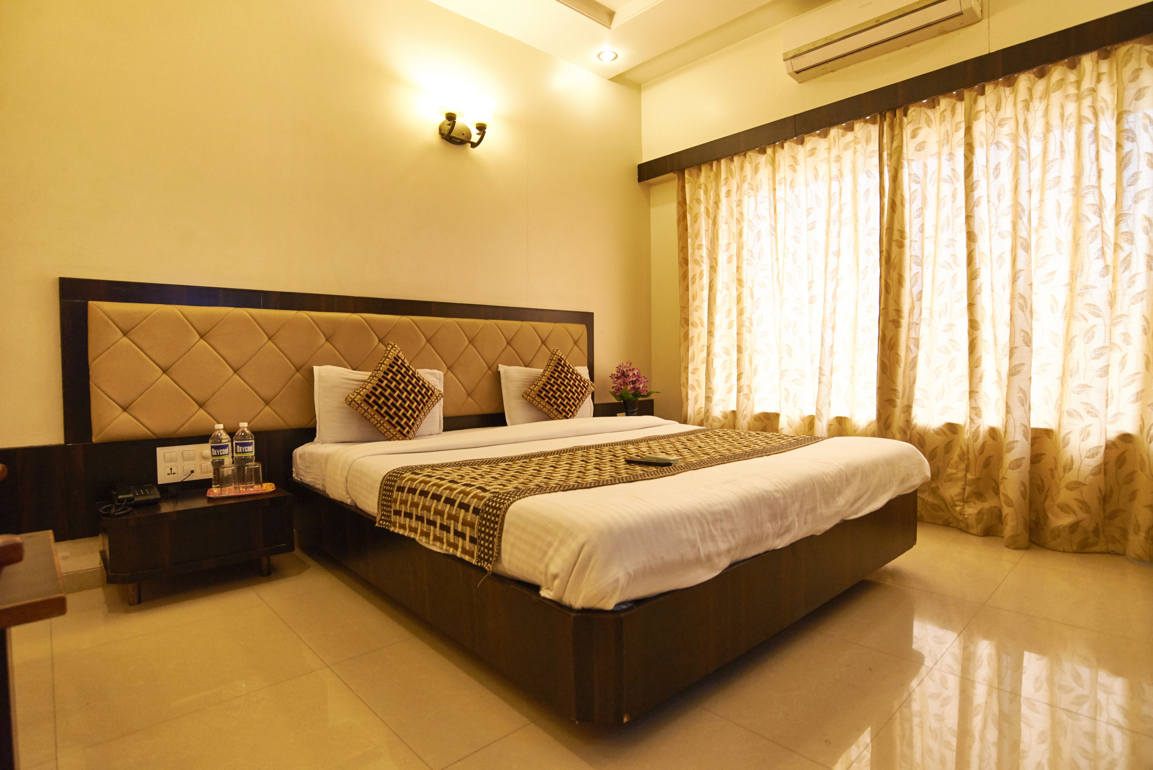 A hotel room with a large bed, a bedside table with a phone and water bottles, and a window with curtains.