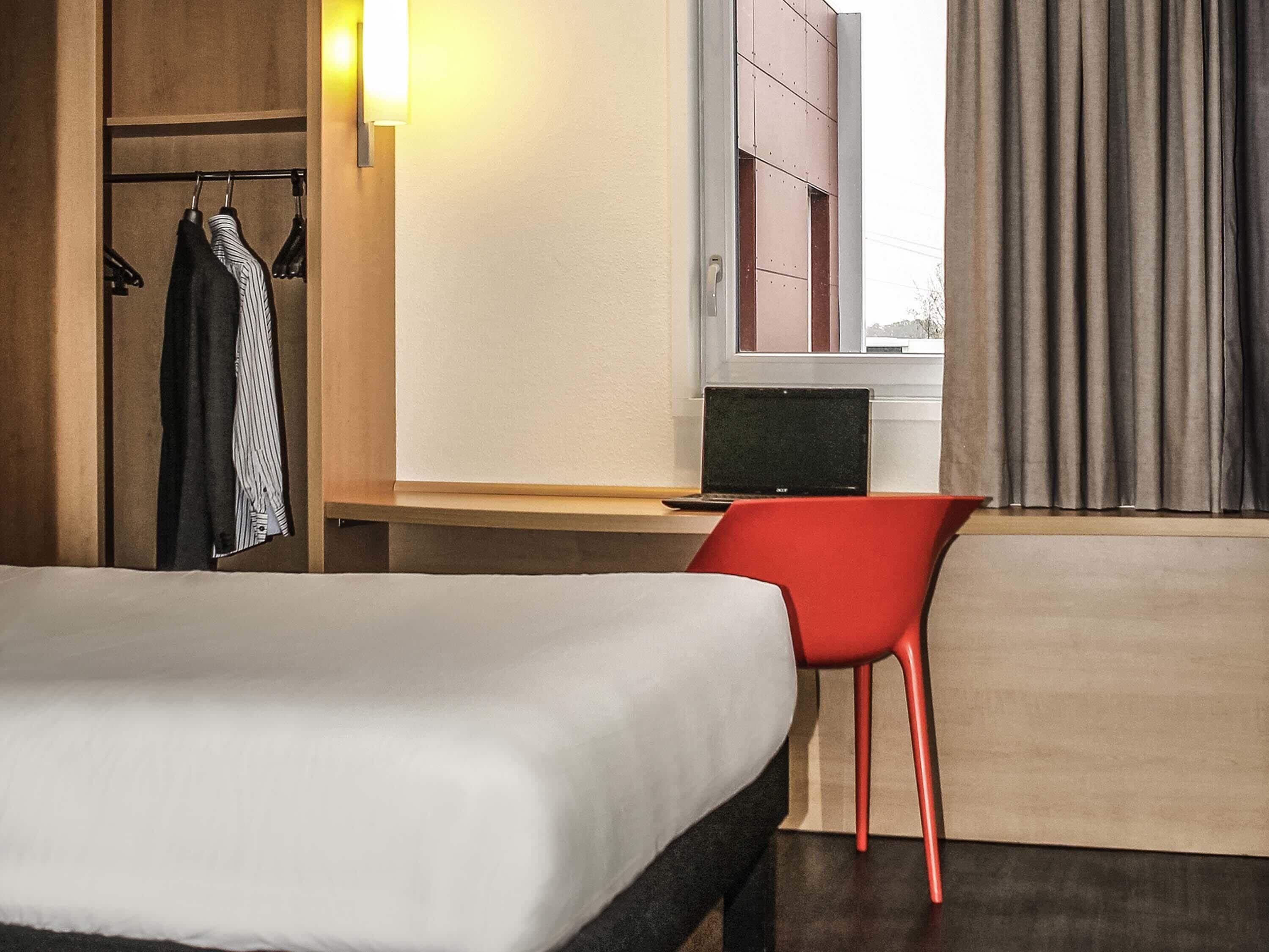 Een hotelkamer met een bed, een bureau met een laptop, een rode stoel, een kledingkast en een raam met gordijnen.