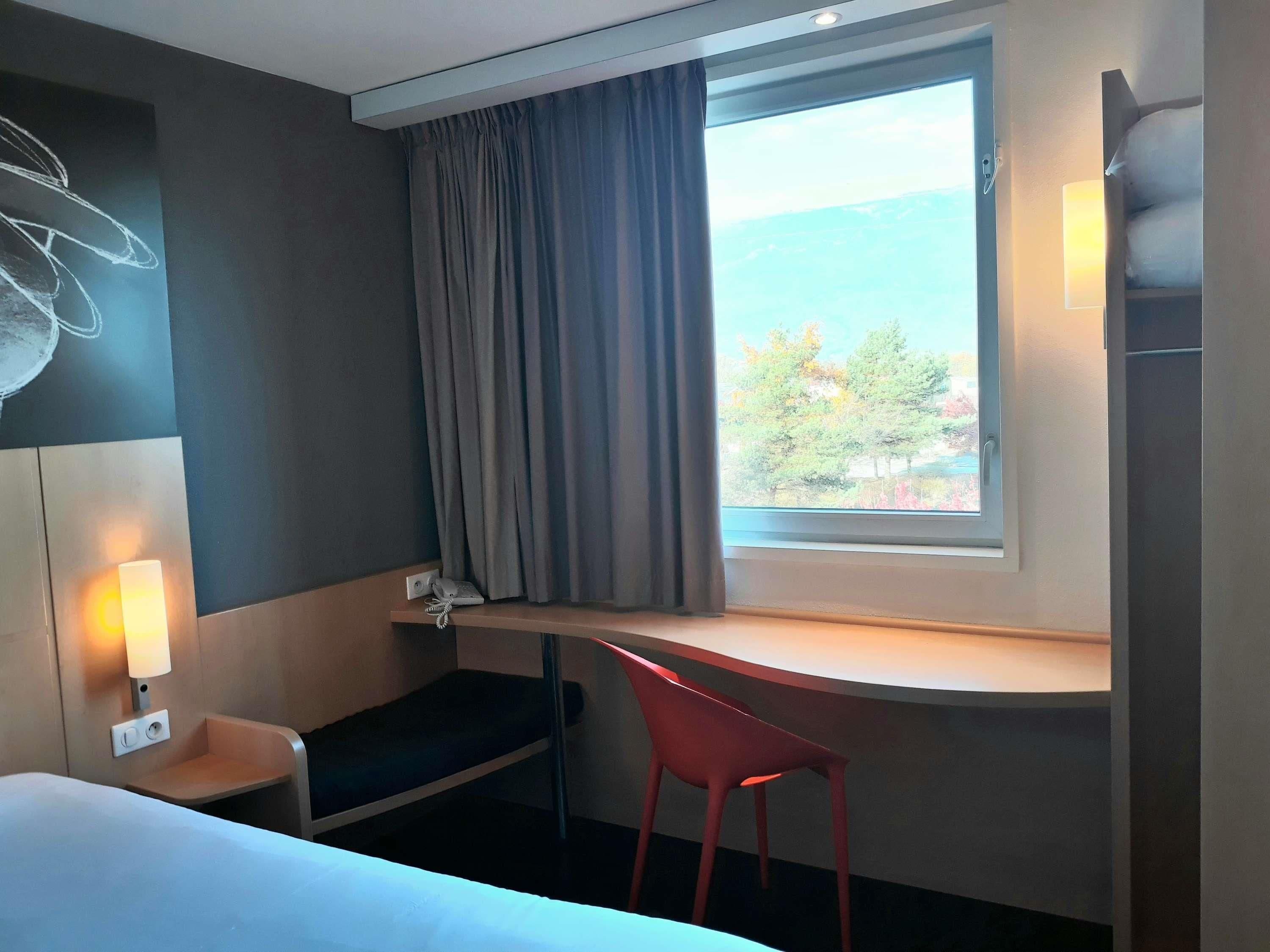 A hotel room with a bed, a desk, a chair, a window with a view, and a wall-mounted lamp.
