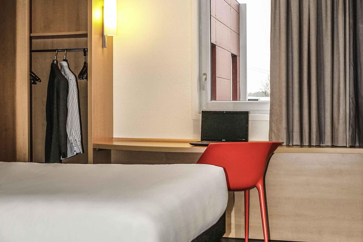 Habitación de hotel con cama, escritorio con computadora portátil, silla roja, armario y ventana con cortinas.