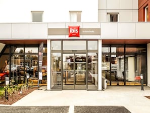 A modern hotel entrance with glass doors, a sign for 'ibis Hotels St Genis Pouilly,' and a small garden area with plants and a bench.