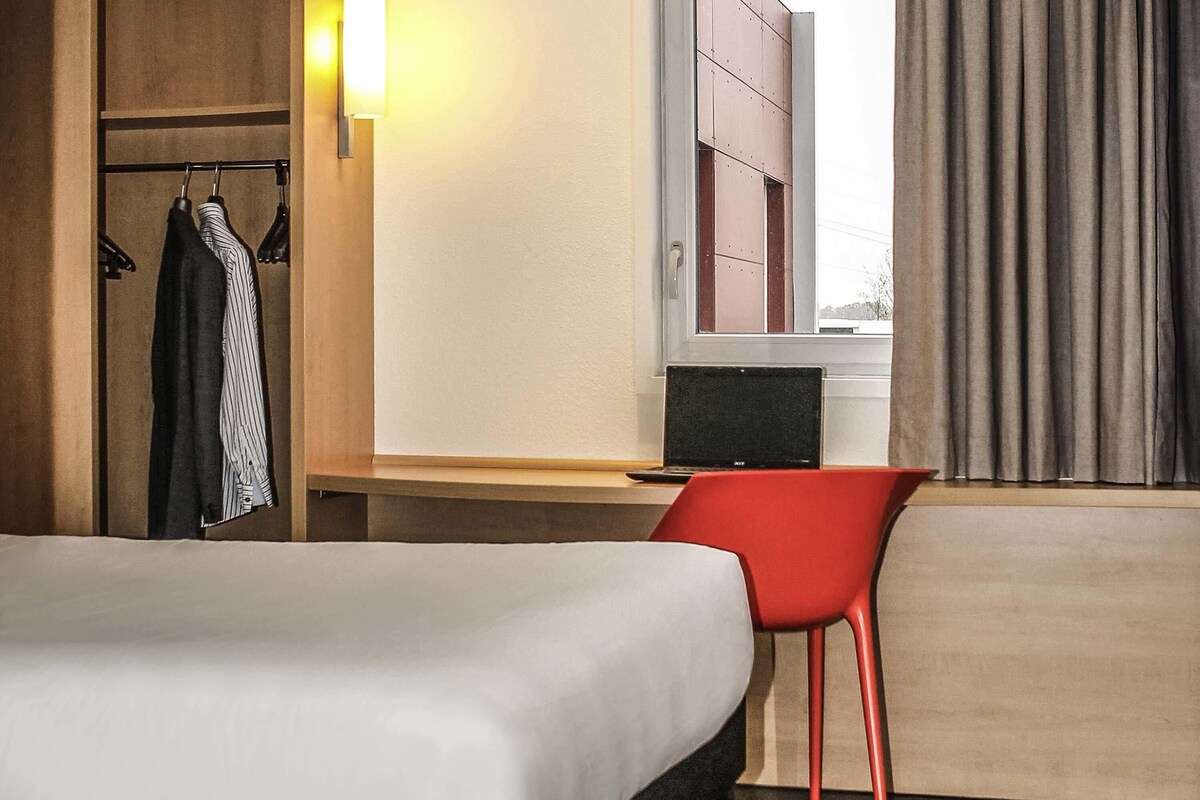 A hotel room with a bed, a desk with a laptop, a red chair, a wardrobe, and a window with curtains.